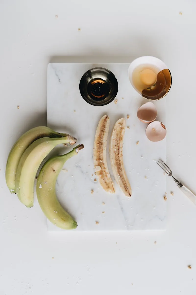 Vista superior de la composición de plátanos maduros cortados y un tazón de huevo crudo sobre una tabla de mármol blanco decorada con cáscara de huevo rota y tenedor